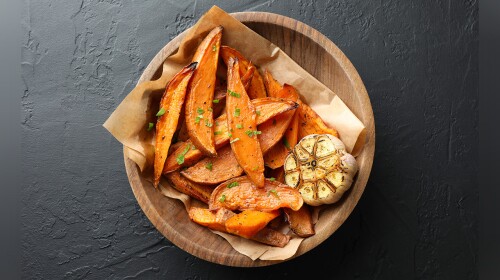 Overhead view of cooked sweet potatoes in a wooden bowl with parsley, a head of roasted garlic and parchment paper in a wooden bowl on a gray slate table.